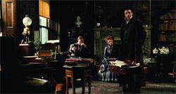 A man stands near a desk while a man and woman sit in a dark, antique-style room.
