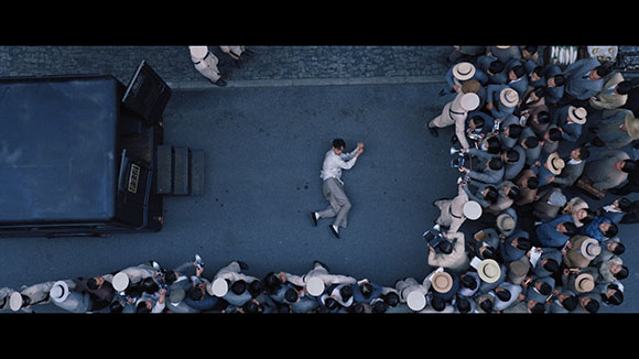 Aerial view of a man lying on a paved surface between a dark vehicle and a large crowd of people.