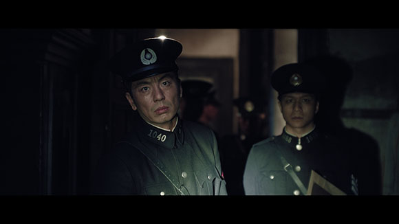 Two men in dark military uniforms and peaked caps stand in a dark, shadowy hallway.
