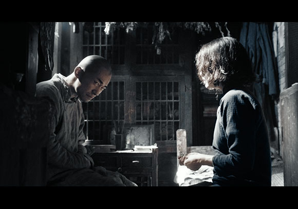 A bald man sits facing a young person with dark hair in a dark, wooden interior.