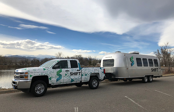 A white pickup truck with teal graphics towing a silver Airstream trailer parked near a lake.