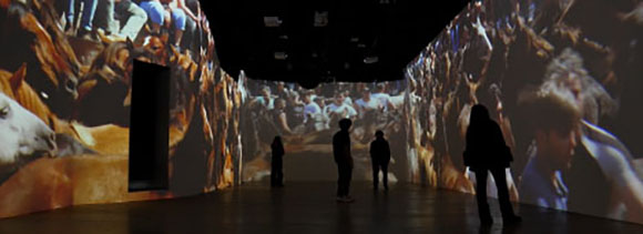 Silhouetted people walk through a dark gallery featuring large-scale video projections on the walls.