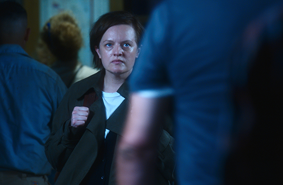 A woman with short dark hair looks forward with a serious expression in a dimly lit room.