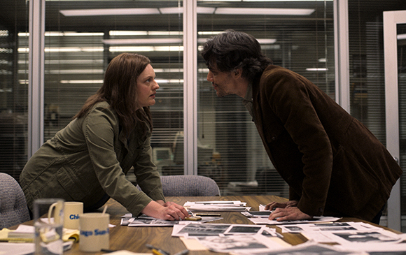 A woman in a green jacket and a man in a brown jacket lean over a table, staring intensely at each other.
