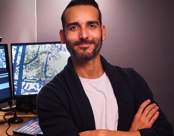 A smiling man with dark hair and a beard stands with arms crossed in front of computer monitors.