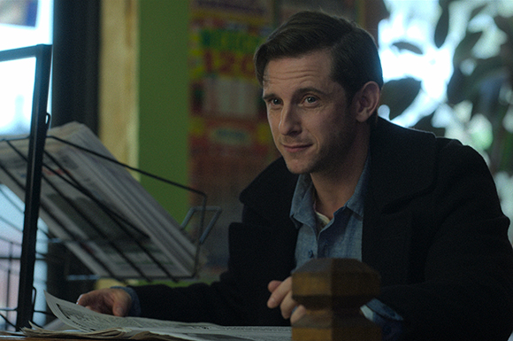 A man in a dark coat and blue shirt sits at a table with newspapers and a wooden object.