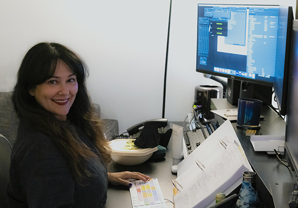 A woman with long dark hair smiles while sitting at a desk with two computer monitors and office supplies.