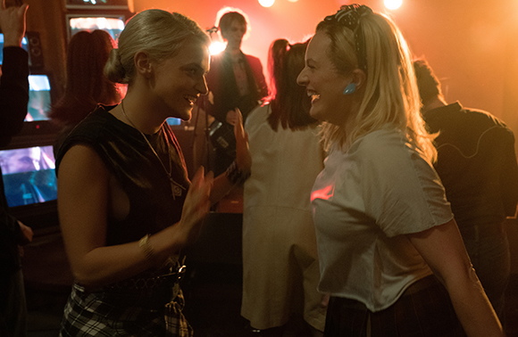 Two blonde women smile and laugh at each other in a crowded, dimly lit room with warm lighting.