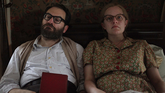 A bearded man in glasses and a woman in a patterned dress sit side by side looking forward.