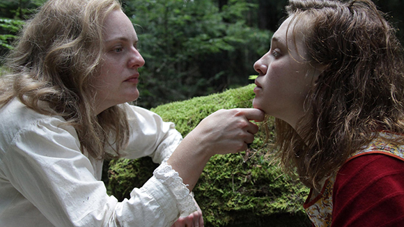 A woman in a white blouse holds the chin of another woman in a red shirt in a wooded area.