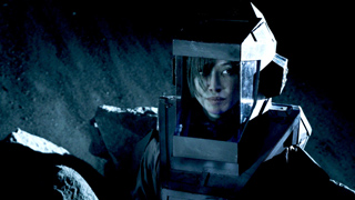 A person wearing a large, metallic, boxy futuristic helmet looking toward the camera in low light.