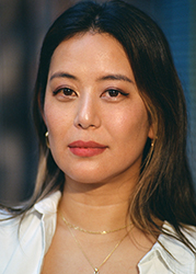 A woman with long dark hair and gold jewelry looks forward against a blurred background.