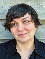A person with short, wavy dark hair and a black collared shirt smiling slightly against a wooden background.