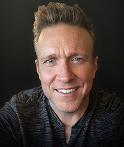 A smiling man with light brown hair looking directly at the camera against a dark background.