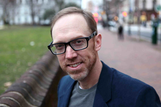 A man with short brown hair and glasses smiles while wearing a navy blazer and grey shirt.