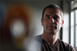 A man with short dark hair looks directly at the camera in a dimly lit room.