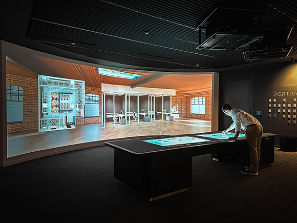 A person leans over a large digital display table in a dark room with a massive wall projection.