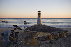 A white lighthouse stands on a rocky islet at sunset near a beach with film production equipment in the foreground.