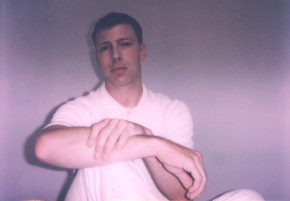 A man wearing a white short-sleeved polo shirt poses with his arms crossed in front of a light purple wall.