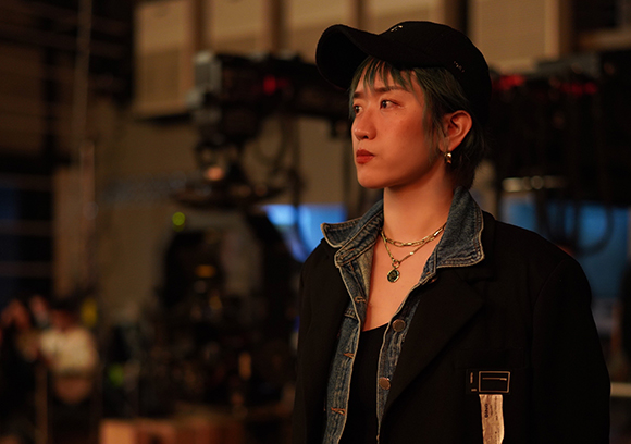 A person wearing a black cap, denim jacket, and gold necklace looking off-camera in a dimly lit studio.