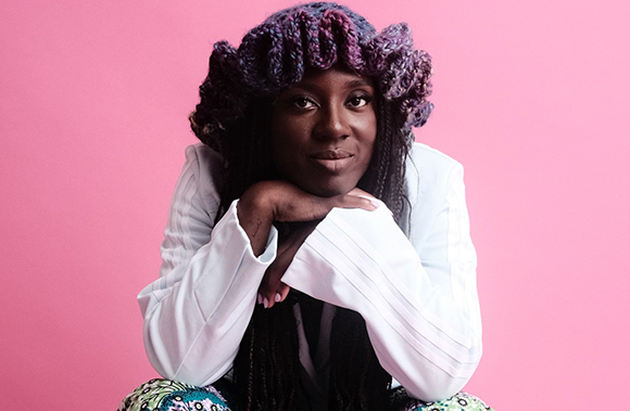 A person with dark skin wearing a voluminous purple textured headpiece and a white blazer against a pink backdrop.