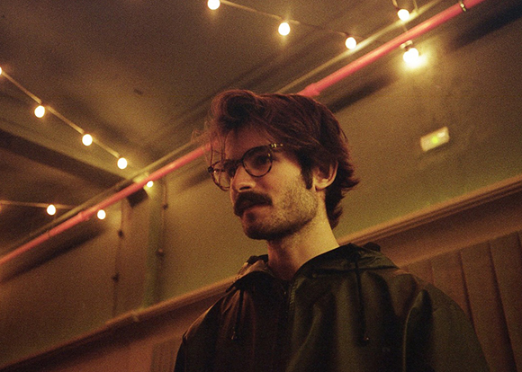 A man with dark hair, glasses, and a mustache looks sideways under warm string lights in a dim room.