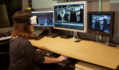 Person wearing a VR headset sits at a desk with three computer monitors displaying digital graphics.