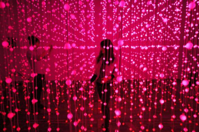 Two people walk through a room filled with hanging pink glowing spheres on vertical strings.