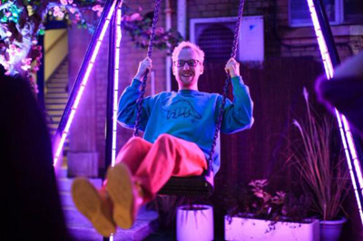 A man with glasses and a blue sweatshirt sits on a swing, smiling under purple neon lights.