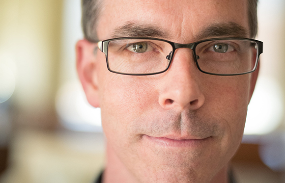 Close-up portrait of a man with short dark hair and black-rimmed glasses looking directly at the camera.