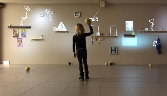 A person in black clothing stands in a room facing a wall decorated with drawings and objects.