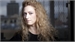 A woman with long, curly blonde hair looks forward against a blurred city background.