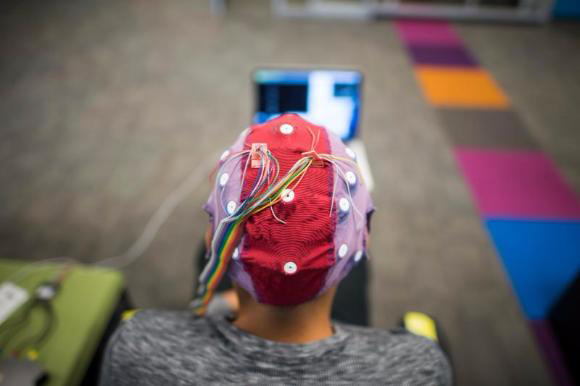 A person from behind wearing a red and purple EEG cap with multicolored wires connected to a laptop.