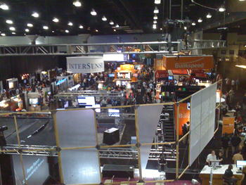 Busy convention hall with numerous booths, displays, and crowds of people. Overhead lighting illuminates the space.