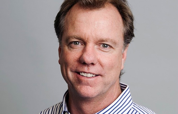 A smiling man with light brown hair wearing a blue and white striped collared shirt against a gray background.