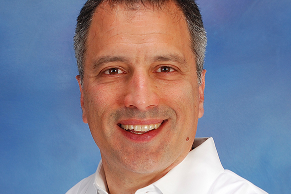 A middle-aged man with short dark hair smiling against a solid blue background.