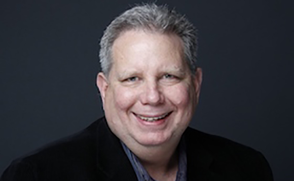 A smiling man with short graying hair wearing a black blazer and dark shirt against a dark background.