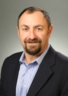 A man with short dark hair and a goatee wearing a light blue shirt and dark blazer.