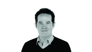 A man with dark hair wearing a dark sweater over a light collared shirt against a white background.