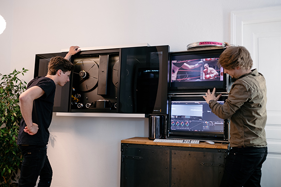 Two men examine a large black media storage unit and two stacked computer monitors on a desk.