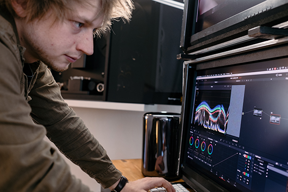 A man with light hair looks intently at a computer monitor displaying video editing software.