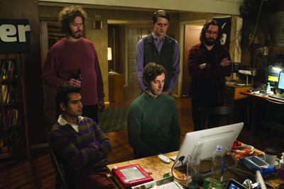Five men stand and sit in a dimly lit, cluttered office filled with books, computers, and equipment.