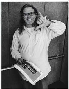 A smiling man with long hair and glasses holds a portfolio and makes an 'L' shape with his hand.