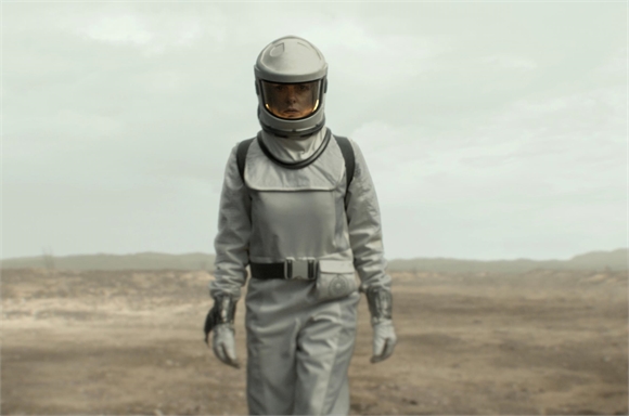 A person wearing a grey astronaut suit and helmet walks across a desolate, flat landscape under an overcast sky.