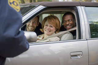 A smiling man, woman, and man sitting in a car while a police officer stands beside them.