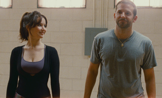 A woman in a black top looks at a man in a grey t-shirt in a room with light brick walls.