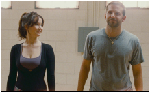 A woman in a black top smiles at a man in a grey t-shirt standing in a room with light brick walls.