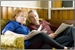 A woman in a blue shirt and a man in a maroon shirt sit on a couch reading magazines.