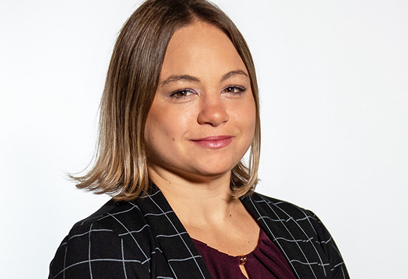 A woman with shoulder-length blonde hair wearing a black grid-patterned blazer and a dark maroon top.