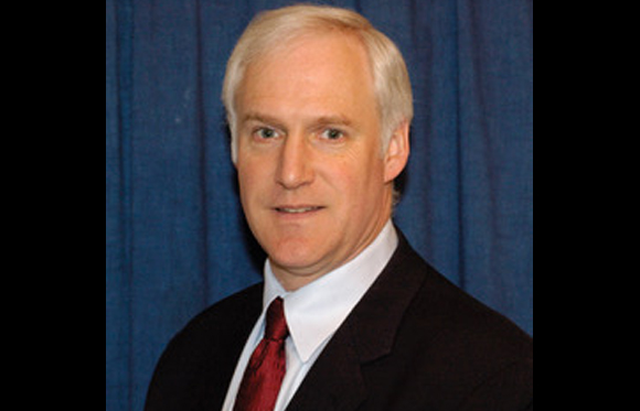 A man with white hair wearing a dark suit, white shirt, and red tie against a blue curtain.